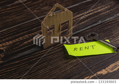 Cardboard house model and key with rent note on wooden surface Cardboard house model and key with rent note on wooden surface 133193603