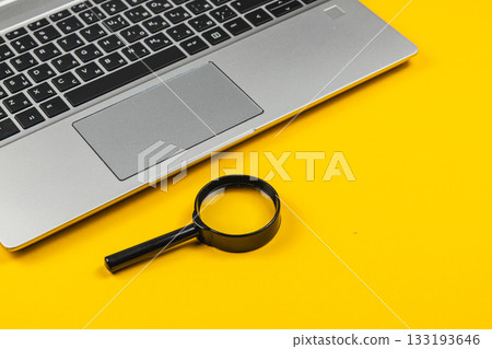 Laptop and magnifying glass on bright yellow surface for clear visuals Laptop and magnifying glass on bright yellow surface for clear visuals 133193646