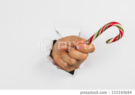 Hand holding a candy cane through a torn hole in a white background Hand holding a candy cane through a torn hole in a white background 133193684