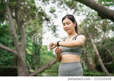 Young woman monitoring her fitness progress on smartwatch in a serene outdoor park surrounded by lush trees and greenery during a sunny day Young woman monitoring her fitness progress on smartwatch in a serene outdoor park surrounded by lush trees and greenery during a sunny day 133193936