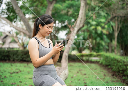 Young woman in sportswear sitting outdoors, using smartphone in nature, practicing wellness, enjoying technology, engaged in physical activity, fitness lifestyle 133193938
