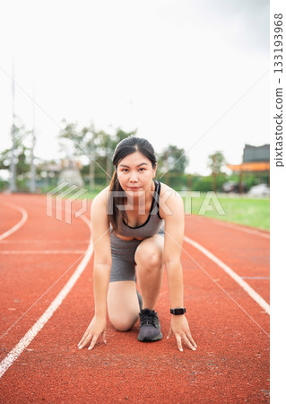 Motivated Female Athlete Preparing for Race on Outdoor Track in Sportswear, Focused Expression with Natural Scenery in Background 133193968