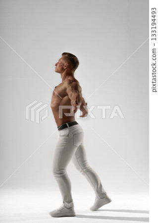 athletic man stands in a studio with a confident posture showing off his muscular physique. He is dressed in fitted pants and sports shoes. The backdrop is minimalistic emphasizing his form. athletic man stands in a studio with a confident posture showing off his muscular physique. He is dressed in fitted pants and sports shoes. The backdrop is minimalistic emphasizing his form. 133194493