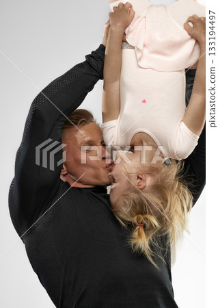 father lifts his blonde daughter upside down sharing a playful kiss. The little girl wears a light pink outfit showcasing their close bond and happiness together. father lifts his blonde daughter upside down sharing a playful kiss. The little girl wears a light pink outfit showcasing their close bond and happiness together. 133194497