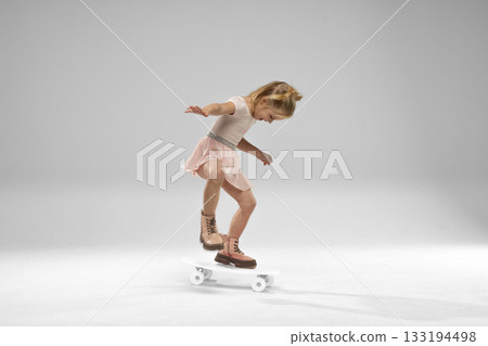 A small blonde girl stands on a skateboard in a studio. She balances carefully wearing a cute outfit and focused on her activity. Bright lighting highlights her joy while skating. A small blonde girl stands on a skateboard in a studio. She balances carefully wearing a cute outfit and focused on her activity. Bright lighting highlights her joy while skating. 133194498