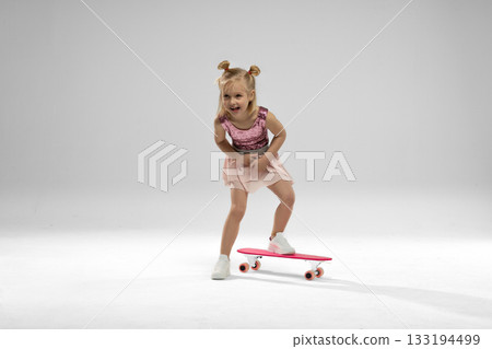 small blonde girl rides a pink skateboard in a bright studio. She smiles with excitement while balancing and showing her skills in this playful moment. 133194499