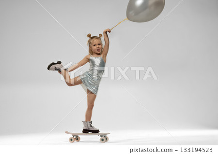 small blonde girl confidently rides a skateboard. She balances on one leg while holding a balloon dressed in a shiny outfit that adds to her playful charm. small blonde girl confidently rides a skateboard. She balances on one leg while holding a balloon dressed in a shiny outfit that adds to her playful charm. 133194523