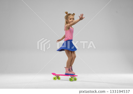 small blonde girl is having fun in a studio skillfully balancing on a pink skateboard. She wears a pink top blue skirt and playful boots showcasing her joy and energy. 133194537