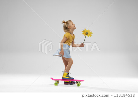 small blonde girl is skateboarding in a bright studio holding a yellow flower and smiling happily. She wears a yellow top and a light blue skirt capturing a joyful scene. 133194538