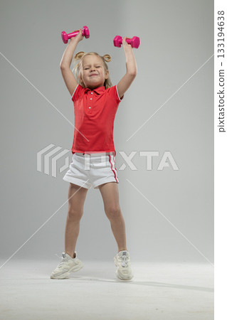 small blonde girl lifts pink dumbbells high above her head. She wears a red shirt and white shorts. The scene captures her joy and enthusiasm for fitness. 133194638