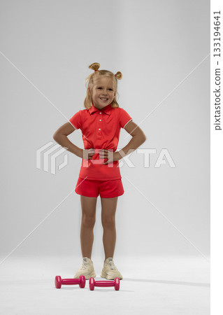 small blonde girl stands confidently with her hands on her hips. She wears a vibrant red outfit and is ready to lift pink dumbbells promoting fitness and fun. small blonde girl stands confidently with her hands on her hips. She wears a vibrant red outfit and is ready to lift pink dumbbells promoting fitness and fun. 133194641