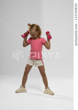 small blonde girl joyfully lifts pink dumbbells. She wears a pink shirt and shorts showing enthusiasm while exercising. 133194650