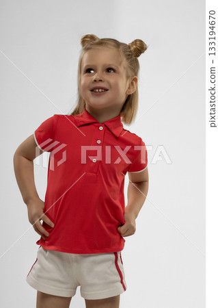 cheerful little girl with blonde hair stands confidently in a studio. She wears a red shirt and white shorts displaying a joyful smile while posing for a picture. cheerful little girl with blonde hair stands confidently in a studio. She wears a red shirt and white shorts displaying a joyful smile while posing for a picture. 133194670