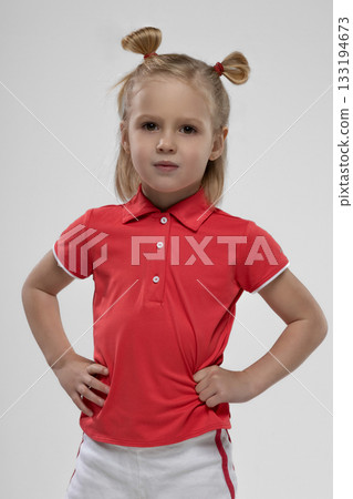 little blonde girl stands confidently with her hands on her hips. She wears a red polo shirt and has her hair styled in playful pigtails showcasing her spirited personality. 133194673