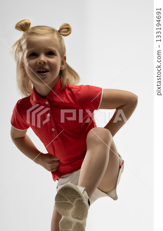 Blonde girl in a red shirt shows a playful pose in a studio. She wears a bright smile while balancing on one leg capturing a moment of joy and creativity. Blonde girl in a red shirt shows a playful pose in a studio. She wears a bright smile while balancing on one leg capturing a moment of joy and creativity. 133194691