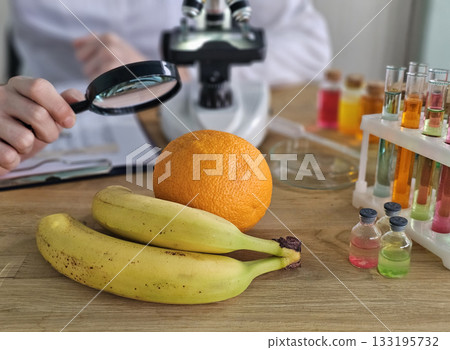 Scientist examining fruits with magnifying glass near colorful liquids and microscope in a lab Scientist examining fruits with magnifying glass near colorful liquids and microscope in a lab 133195732