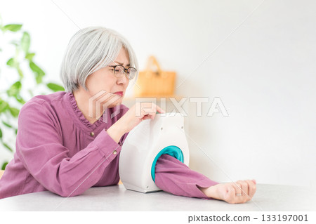 A gray-haired senior woman measuring her blood pressure using a blood pressure monitor in her living room at home A gray-haired senior woman measuring her blood pressure using a blood pressure monitor in her living room at home 133197001