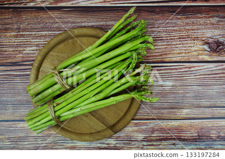 Bunch of fresh green asparagus stems with cutting wooden board on wooden background Bunch of fresh green asparagus stems with cutting wooden board on wooden background 133197284