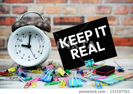 Keep It Real text on paper card with alarm clock and stationary on wooden background Keep It Real text on paper card with alarm clock and stationary on wooden background 133197561