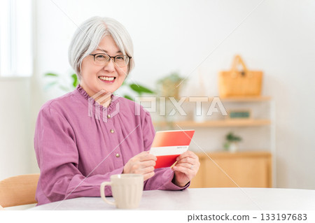 Smiling senior woman looking at her savings passbook in the living room of her home (retirement funds, asset management, deposits, income, living expenses) 133197683