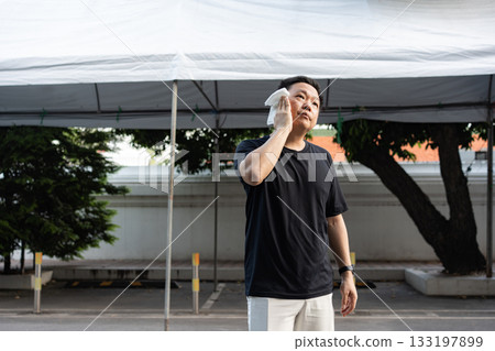Cooling Off the Heat, a man wipes sweat from his face under a shade capturing the relief and calmness after facing the midday warmth. 133197899