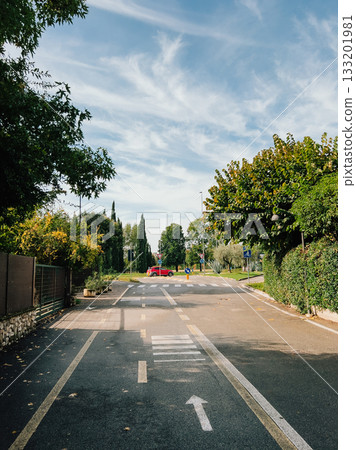 Paved suburban street featuring bike lane and pedestrian crossing, lined with trees and hedges, showcasing parked red car under blue sky with clouds. Residential street with bike lane, green landscape Paved suburban street featuring bike lane and pedestrian crossing, lined with trees and hedges, showcasing parked red car under blue sky with clouds. Residential street with bike lane, green landscape 133201981