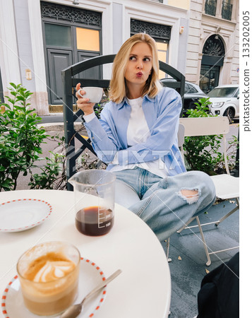 Young blonde woman is sitting at table in cafe, enjoying coffee break and making funny face while holding cup of coffee. Woman sitting at cafe table with coffee and making playful expression 133202005