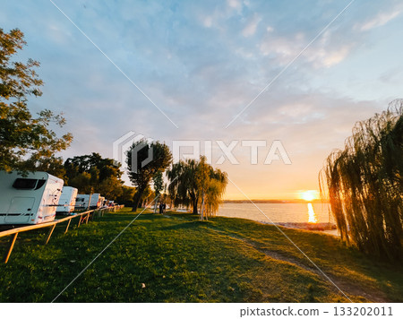 Campers enjoying warm glow of golden hour light during sunset, gathered near tranquil lake with recreational vehicles parked on lush grassy field, embracing nature beauty. Camp, recreational vehicles 133202011