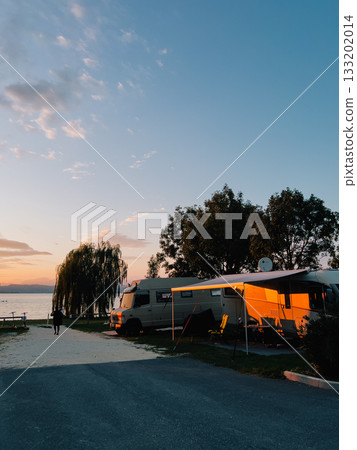 Campers parked by tranquil lake during sunset, offering serene and picturesque backdrop for unforgettable camping experience filled with relaxation and adventure. Recreational vehicles near shoreline 133202014