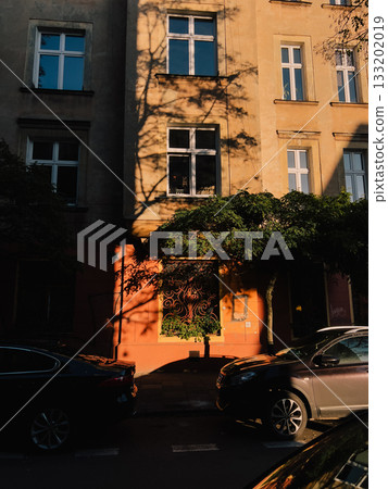 Warm sunlight illuminating colorful building facade with parked cars and casting tree shadows on wall, creating picturesque urban scene. Warm sunlight casting tree shadows on colorful building facade 133202019