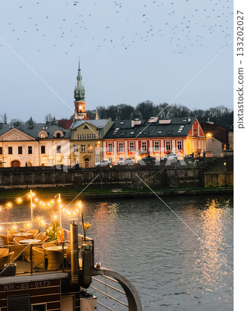 Birds flying over Krakow riverside with illuminated restaurant and St. Andrew Church in background at dusk, creating magical atmosphere. Silhouetted birds soaring above Krakow riverbank. Evening City Birds flying over Krakow riverside with illuminated restaurant and St. Andrew Church in background at dusk, creating magical atmosphere. Silhouetted birds soaring above Krakow riverbank. Evening City 133202027