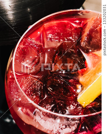 Refreshing red cocktail served in balloon glass, filled with ice cubes and garnished with vibrant lemon wedge, creating inviting and stylish drink perfect for any celebration. Red cocktail with ice 133202031