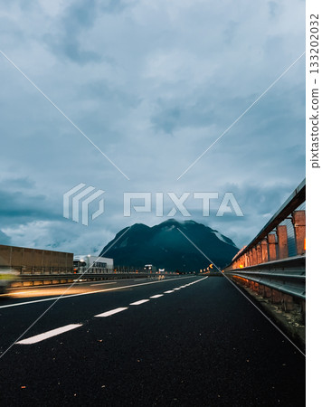 Empty highway disappearing into distance towards mountains under cloudy sky at dusk, with truck driving in distance and lights of passing car creating light trails. Asphalt road, mountain silhouettes 133202032