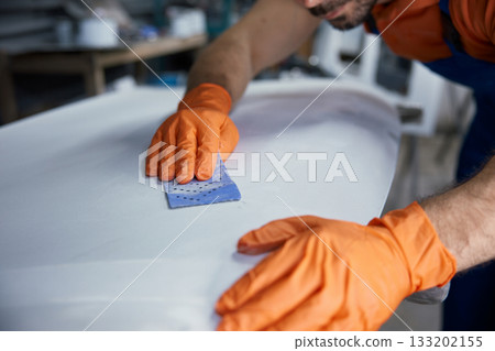 Craftsman sanding surfboard in workshop with hands with orange gloves finishing board 133202155