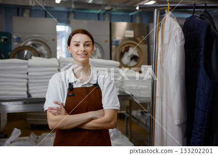 A woman worker in an apron stands with arms crossed in a laundromat 133202201