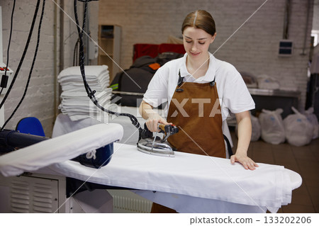 Woman worker wearing an apron is skillfully ironing a shirt on a table 133202206