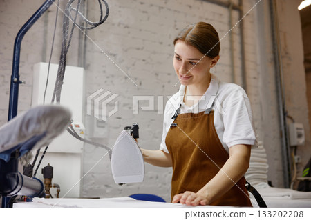 A woman worker wearing an apron is skillfully ironing a shirt on a table A woman worker wearing an apron is skillfully ironing a shirt on a table 133202208