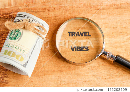 A stack of one hundred dollar bills secured with twine sits beside a magnifying glass that shows the words TRAVEL VIBES on a textured wooden surface 133202309