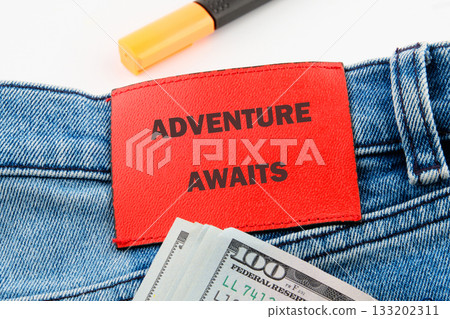 A close up features blue jeans with a red patch saying Adventure Awaits. A stack of United States currency and a marker are visible A close up features blue jeans with a red patch saying Adventure Awaits. A stack of United States currency and a marker are visible 133202311