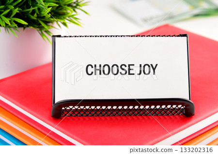 A close-up shows a desk with a sign reading CHOOSE JOY resting on colorful books. A potted plant sits nearby. It is a simple reminder to find happiness in every moment 133202316