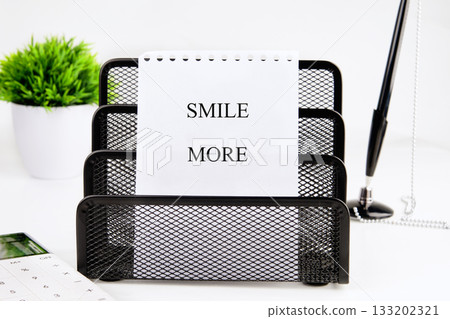 A black wire organizer holds a notepad with the words SMILE MORE written on it. A pen, calculator, and plant sit nearby on a white desk, creating a bright and encouraging workspace 133202321