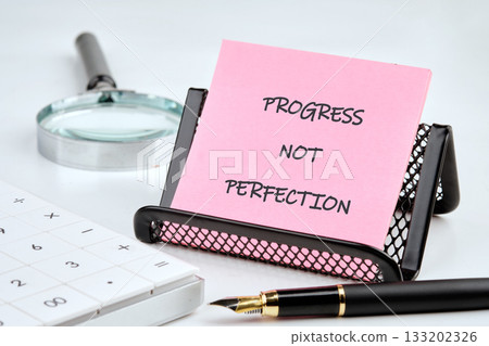 Pink notepad displays the words PROGRESS NOT PERFECTION. Calculator, pen, and magnifying glass are nearby. The scene speaks of improvement, focus, and striving forward Pink notepad displays the words PROGRESS NOT PERFECTION. Calculator, pen, and magnifying glass are nearby. The scene speaks of improvement, focus, and striving forward 133202326