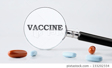 A magnifying glass highlights the word Vaccine on a white background. Several pills, in brown and blue colors, are scattered near the magnifying glass 133202334