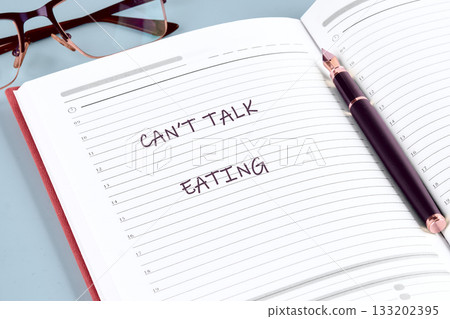Open notebook with the words Cant Talk Eating hand-written on a lined page. A pen rests on the book with glasses in the background on the table Open notebook with the words Cant Talk Eating hand-written on a lined page. A pen rests on the book with glasses in the background on the table 133202395
