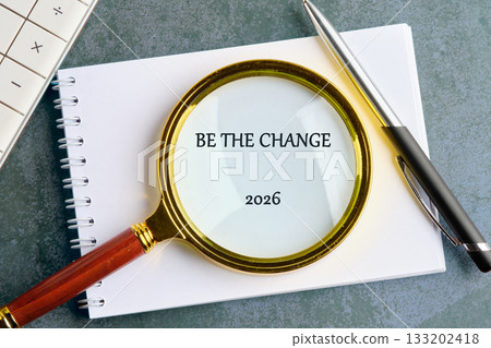 A magnifying glass with a gold rim sits on a notebook with the words BE THE CHANGE and 2026 visible. A pen rests to the side, next to a calculator 133202418
