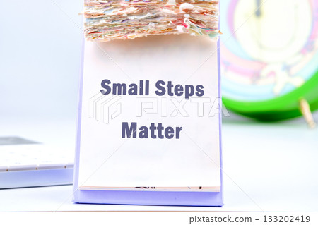 A desk calendar sits open, displaying the motivational message Small Steps Matter, while nearby, a calculator and a clock suggest a focus on time and progress A desk calendar sits open, displaying the motivational message Small Steps Matter, while nearby, a calculator and a clock suggest a focus on time and progress 133202419