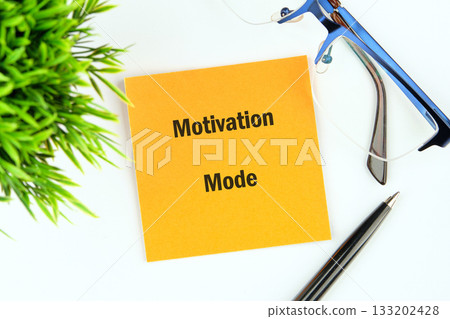 A bright yellow note declares Motivation Mode alongside a sleek pen, blue glasses, and green plant on a crisp white surface, inviting productivity 133202428