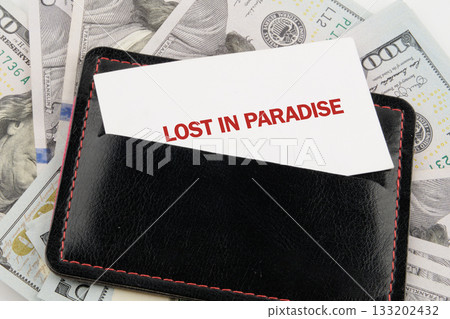 A black leather wallet is nestled among stacks of United States one hundred dollar bills. A card reading LOST IN PARADISE is sticking out of the wallets pocket 133202432
