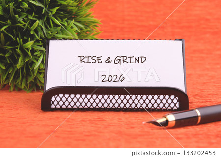 A card with RISE GRIND 2026 sits on an office desk with a fountain pen and a small plant next to it, signaling ambition for the year Twenty Twenty-Six 133202453