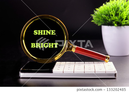 A magnifying glass showcases the words shine bright against a dark background. It sits beside a sleek calculator and a small potted plant, capturing an inspiring moment of clarity A magnifying glass showcases the words shine bright against a dark background. It sits beside a sleek calculator and a small potted plant, capturing an inspiring moment of clarity 133202485